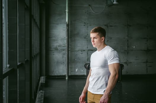 Young sexy men bodybuilder athlete,studio portrait in loft on the background of stylized wall and Black leather sofa, guy model in white T-shirt and brown trousers against background of larger window