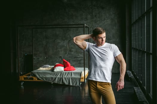 Young sexy men bodybuilder athlete, studio portrait in loft on the background of stylized wall and brutal bed, guy model in white T-shirt and brown trousers against the background of a larger window