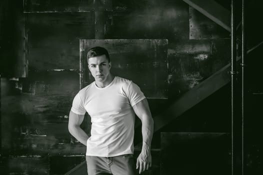 Young sexy men bodybuilder athlete, Studio portrait in a loft on the background of a stylized metal wall, a guy model in a white T-shirt and brown trousers