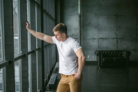 Young sexy men bodybuilder athlete,studio portrait in loft on the background of stylized wall and Black leather sofa, guy model in white T-shirt and brown trousers against background of larger window