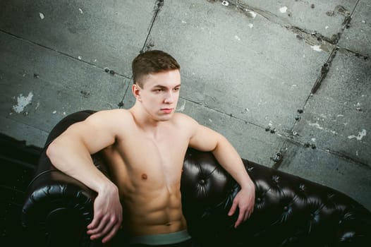 Young sexy men bodybuilder athlete sitting on a black leather couch with a naked torso, studio portrait