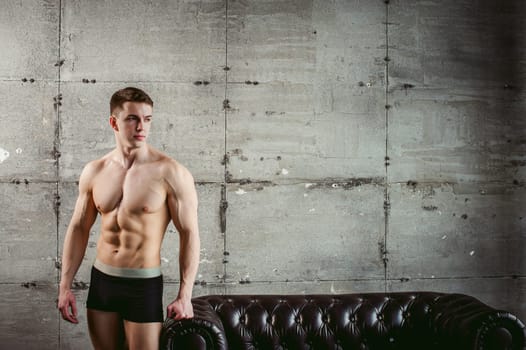 Young sexy men bodybuilder athlete stands at black leather couch with a naked torso, studio portrait