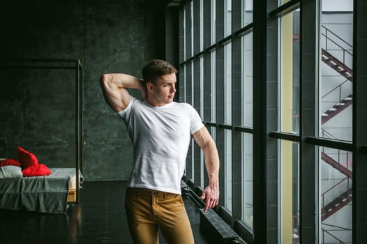 Young sexy men bodybuilder athlete, studio portrait in loft on the background of stylized wall and brutal bed, guy model in white T-shirt and brown trousers against the background of a larger window
