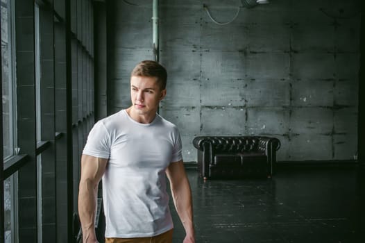 Young sexy men bodybuilder athlete,studio portrait in loft on the background of stylized wall and Black leather sofa, guy model in white T-shirt and brown trousers against background of larger window