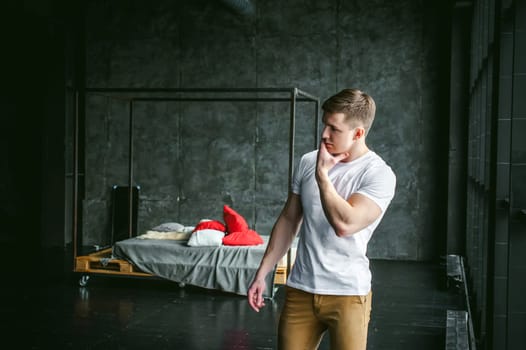 Young sexy men bodybuilder athlete, studio portrait in loft on the background of stylized wall and brutal bed, guy model in white T-shirt and brown trousers against the background of a larger window