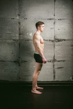 Young sexy male bodybuilder sportsman with a bare torso against a gray wall background, studio portrait of a light-skinned strong man in full growth
