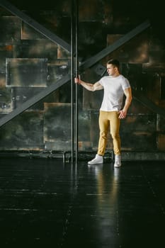 Young sexy men bodybuilder athlete, Studio portrait in a loft on the background of a stylized metal wall, a guy model in a white T-shirt and brown trousers