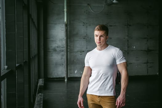 Young sexy men bodybuilder athlete,studio portrait in loft on the background of stylized wall and Black leather sofa, guy model in white T-shirt and brown trousers against background of larger window