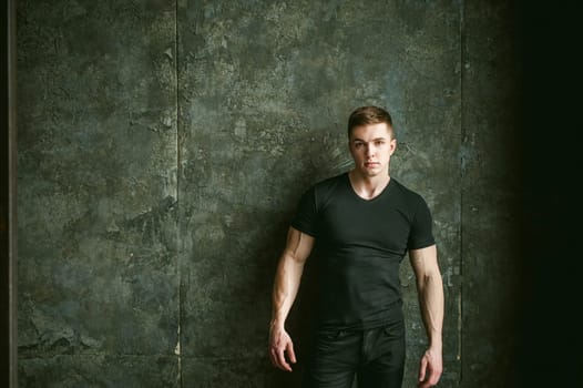 Young sexy men bodybuilder athlete, studio portrait loft on background of stylized Concrete brutal wall, guy model black Tshirt and trousers With powerful muscular arms
