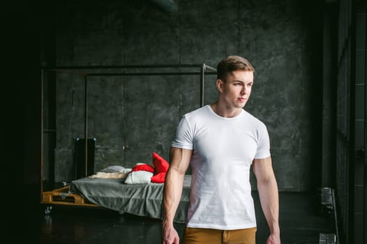 Young sexy men bodybuilder athlete, studio portrait in loft on the background of stylized wall and brutal bed, guy model in white T-shirt and brown trousers against the background of a larger window