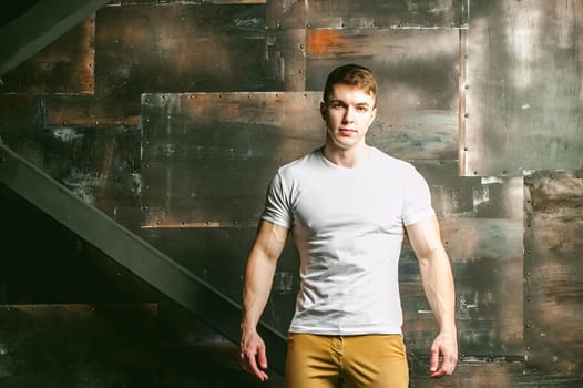 Young sexy men bodybuilder athlete, Studio portrait in a loft on the background of a stylized metal wall, a guy model in a white T-shirt and brown trousers