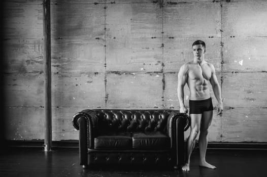 Young sexy men bodybuilder athlete stands at black leather couch with a naked torso, studio portrait