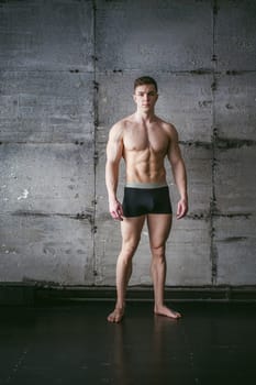 Young sexy male bodybuilder sportsman with a bare torso against a gray wall background, studio portrait of a light-skinned strong man in full growth