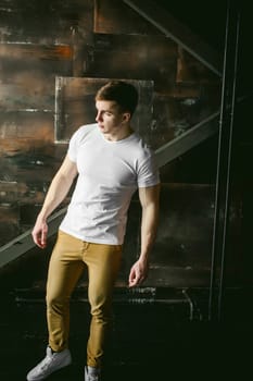 Young sexy men bodybuilder athlete, Studio portrait in a loft on the background of a stylized metal wall, a guy model in a white T-shirt and brown trousers