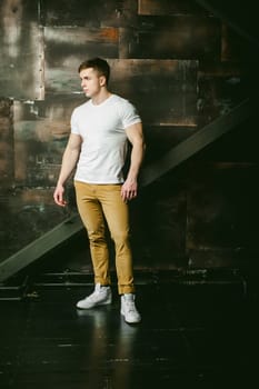 Young sexy men bodybuilder athlete, Studio portrait in a loft on the background of a stylized metal wall, a guy model in a white T-shirt and brown trousers