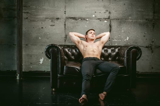 Young sexy men bodybuilder athlete sitting on a black leather couch with a naked torso and black trousers, Threw one's hands behind his head, studio portrait