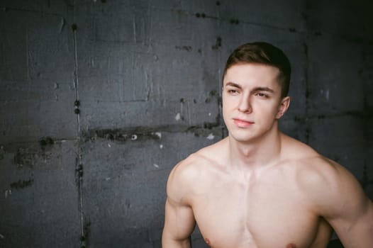 Young sexy male bodybuilder athlete with bare torso against a gray brutal concrete wall, studio portrait of a light-skinned strong man