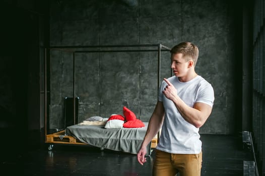 Young sexy men bodybuilder athlete, studio portrait in loft on the background of stylized wall and brutal bed, guy model in white T-shirt and brown trousers against the background of a larger window