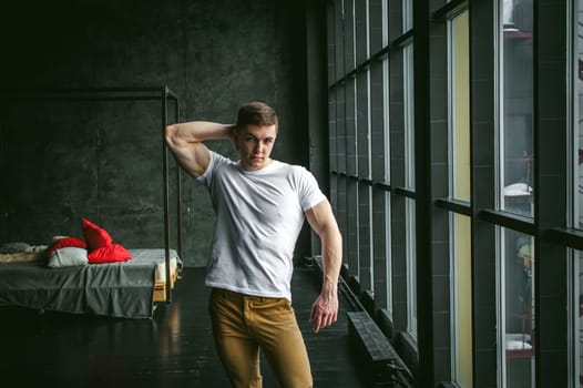 Young sexy men bodybuilder athlete, studio portrait in loft on the background of stylized wall and brutal bed, guy model in white T-shirt and brown trousers against the background of a larger window