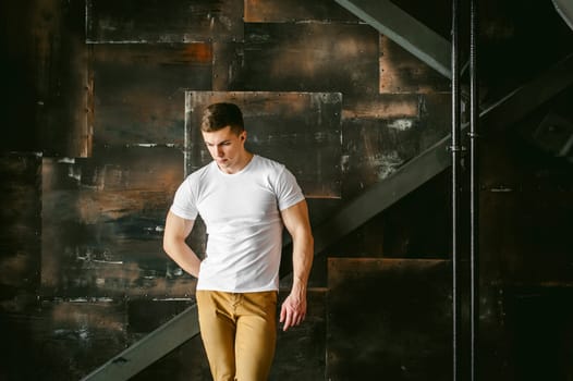 Young sexy men bodybuilder athlete, Studio portrait in a loft on the background of a stylized metal wall, a guy model in a white T-shirt and brown trousers