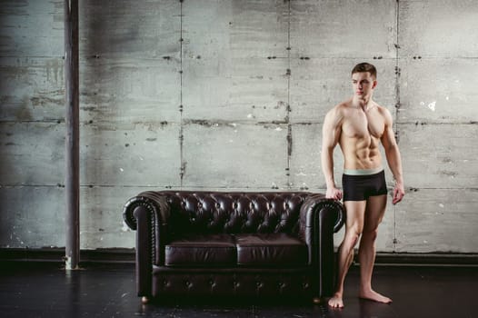 Young sexy men bodybuilder athlete stands at black leather couch with a naked torso, studio portrait