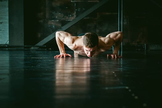 Young sexy male bodybuilder athlete with a bare torso. portrait of a studio of a light-skinned strong man engaged in push-ups, on the floor, straining powerful hands