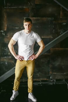 Young sexy men bodybuilder athlete, Studio portrait in a loft on the background of a stylized metal wall, a guy model in a white T-shirt and brown trousers