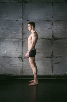 Young sexy male bodybuilder sportsman with a bare torso against a gray wall background, studio portrait of a light-skinned strong man in full growth