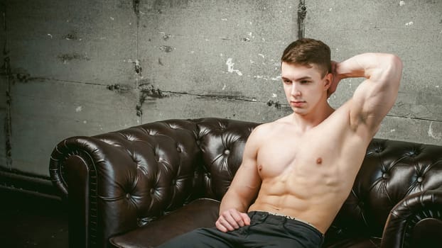 Young sexy men bodybuilder athlete sitting on a black leather couch with a naked torso and black trousers, Threw one's hands behind his head, studio portrait