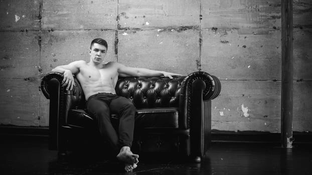 Young sexy men bodybuilder athlete sitting on a black leather couch with a naked torso and black trousers, studio portrait