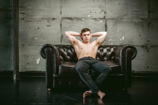 Young sexy men bodybuilder athlete sitting on a black leather couch with a naked torso and black trousers, Threw one's hands behind his head, studio portrait