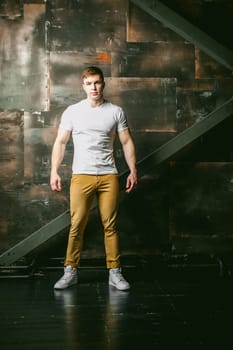 Young sexy men bodybuilder athlete, Studio portrait in a loft on the background of a stylized metal wall, a guy model in a white T-shirt and brown trousers