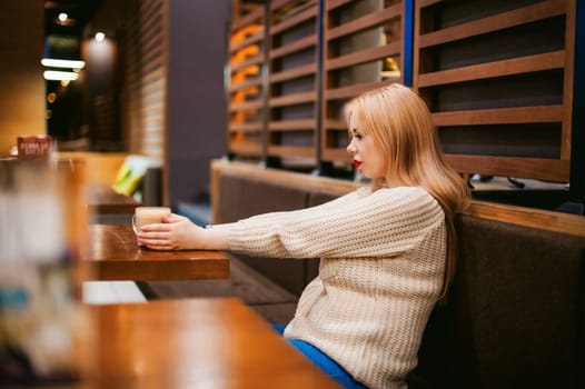 Cute young blonde woman drinks hot coffee from a glass cup in the interior of a coffee house