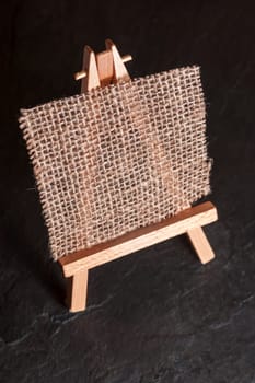 Elevated view of a mini easel with coarse linen fabric as a painting surface against a dark background on slate plate.