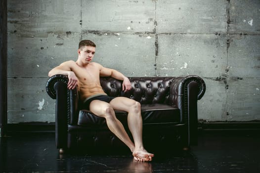 Young sexy men bodybuilder athlete sitting on a black leather couch with a naked torso, studio portrait
