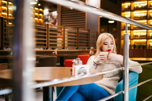 Cute young blonde woman drinks hot coffee from a glass cup in the interior of a coffee house