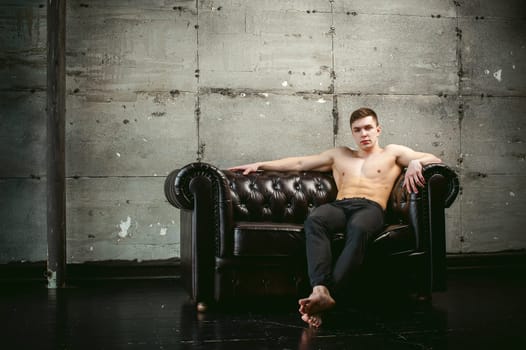 Young sexy men bodybuilder athlete sitting on a black leather couch with a naked torso and black trousers, studio portrait
