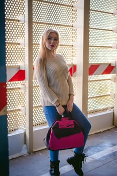 Portrait of a young woman dressed in a knitted sweater and blue jeans, holds in her hand a sports bag. Standing near a metal perforated parking fence construction