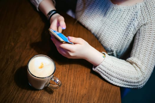 Young blonde woman in a knitted sweater sitting at a table in a cafe with a cup of coffee in her hands, spending time alone, doing internet communication through wifi