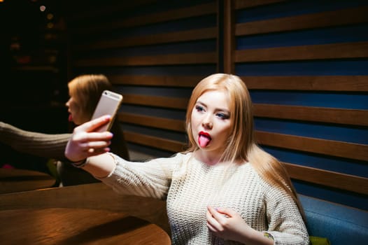 young blond woman in a knitted sweater, making a selfie photo on a smartphone emotionally shows tongue, sitting at a table in a cafe, spending time alone, doing internet communication