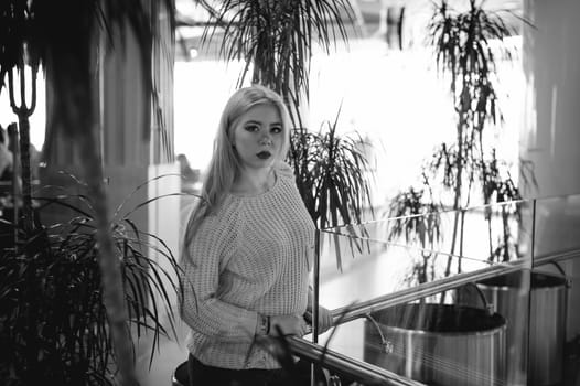 Portrait of a young woman dressed in a knitted sweater, standing near the handrails at a glass fence against the background of the office environment and indoor plant palm trees