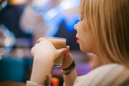 Cute young blonde woman drinks hot coffee from a glass cup in the interior of a coffee house. Abstract blur, out of focus