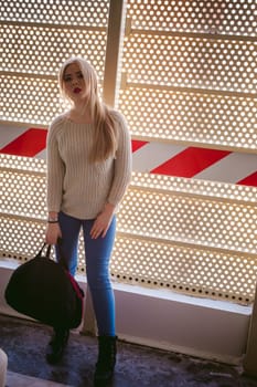 Portrait of a young woman dressed in a knitted sweater and blue jeans, holds in her hand a sports bag. Standing near a metal perforated parking fence construction