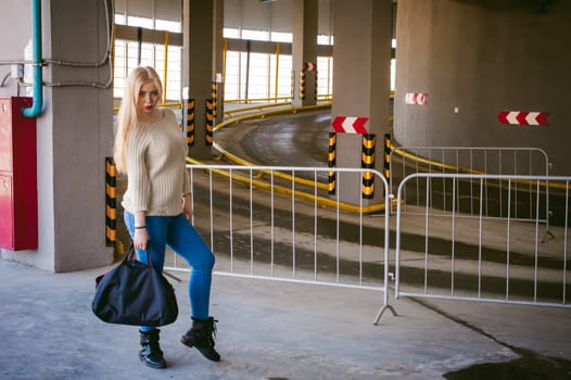 Portrait of a young woman dressed in a knitted sweater and blue jeans, holds in her hand a sports bag. standing on the background of a climb to the parking floor
