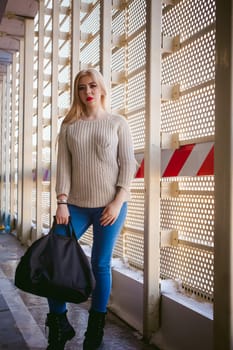 Portrait of a young woman dressed in a knitted sweater and blue jeans, holds in her hand a sports bag. Standing near a metal perforated parking fence construction