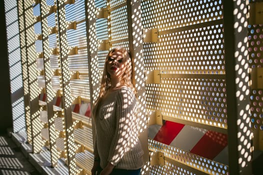 Portrait of a young woman dressed in a knitted sweater and blue jeans. Standing near a metal perforated parking fence construction. Shadows in the form of dots of light fall on her face