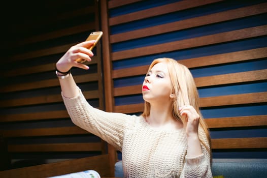 young blond woman in a knitted sweater, making a selfie photo on a smartphone, sitting at a table in a cafe, spending time alone, doing internet communication