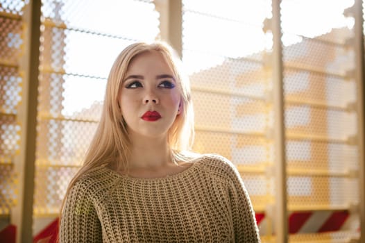 Portrait of a young woman dressed in a knitted sweater and blue jeans. Standing near a metal perforated parking fence construction