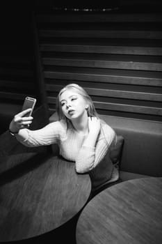 young blond woman in a knitted sweater, making a selfie photo on a smartphone, sitting at a table in a cafe, spending time alone, doing internet communication