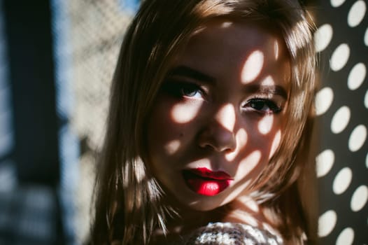 Portrait of a young woman dressed in a knitted sweater and blue jeans. Standing near a metal perforated parking fence construction. Shadows in the form of dots of light fall on her face